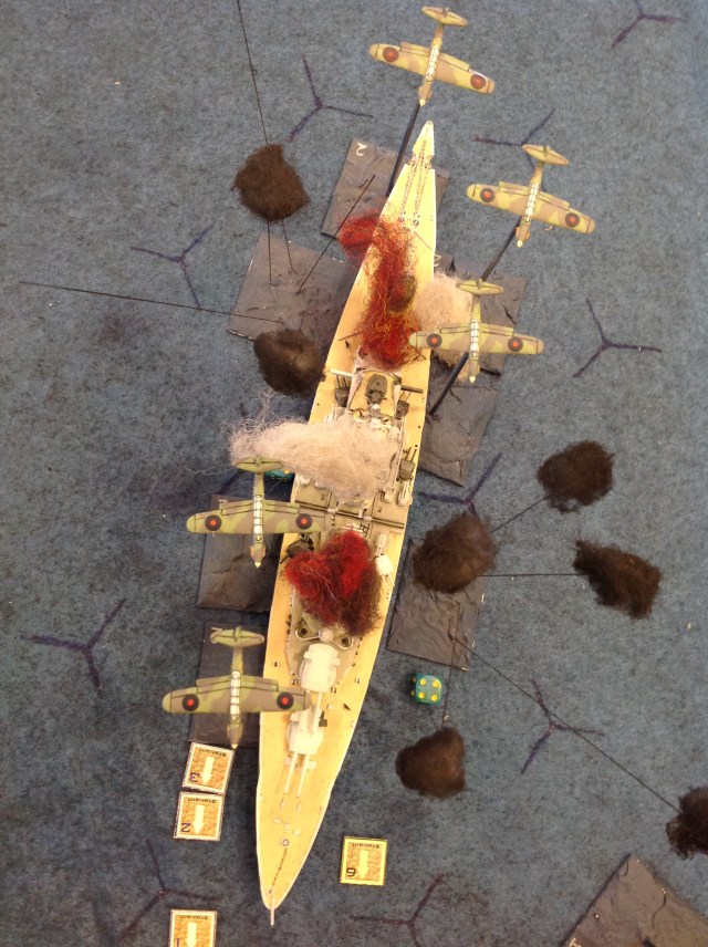 The battleship is wreathed in fire and smoke as the Barracudas recover from their bombing runs.